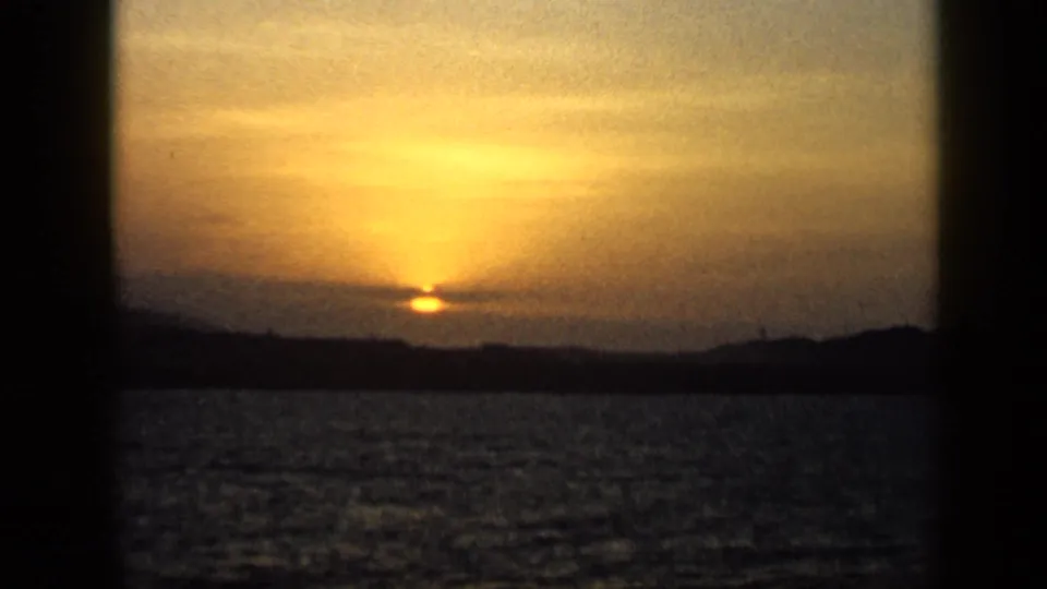 1959 Venezuela 8mm frame showing the sun setting over open water, with a dark coastal horizon and warm orange sky.