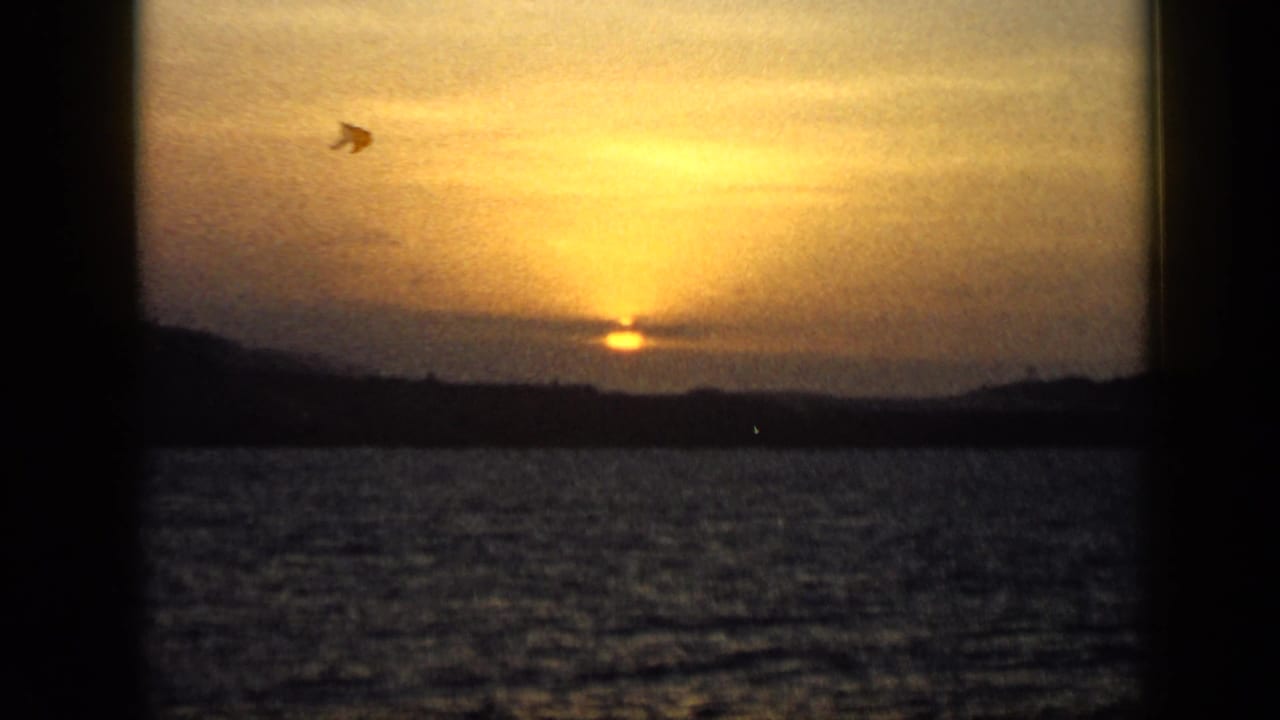 1959 Venezuela 8mm frame showing a low golden sun over water, clouds, and distant dark mountains.