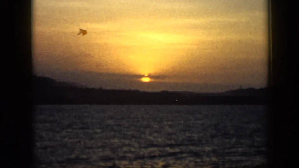 1959 Venezuela 8mm frame showing a low golden sun over water, clouds, and distant dark mountains.