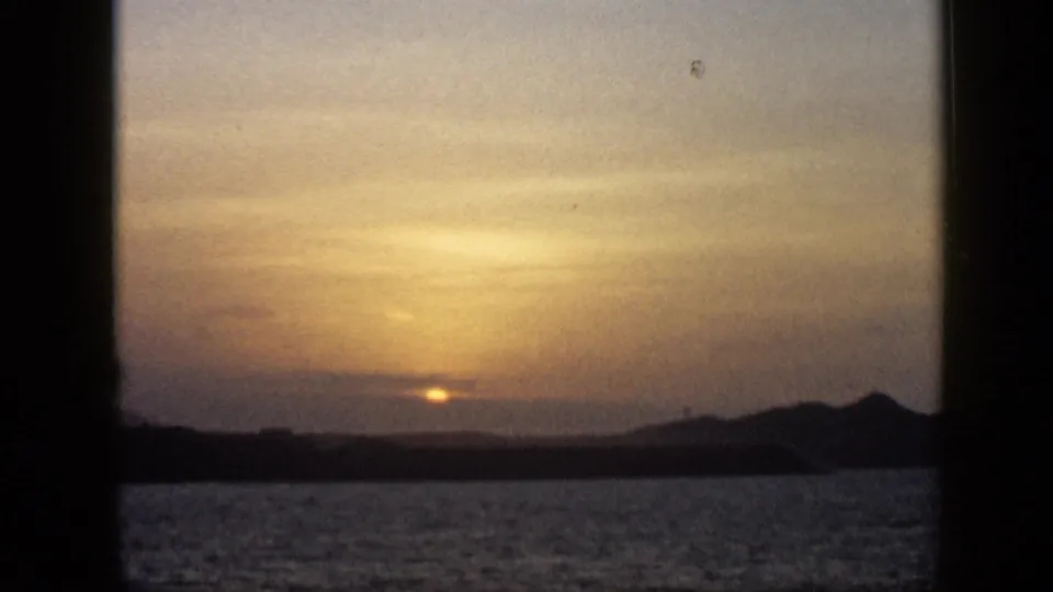 1959 Venezuela 8mm frame with the sun dimmed by cloud cover above a lake or broad waterway and low hills.