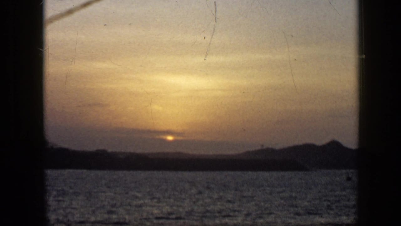 1959 Venezuela 8mm frame with a golden sky, low sun, rippled water, and a dark mountain ridge beyond the sea.