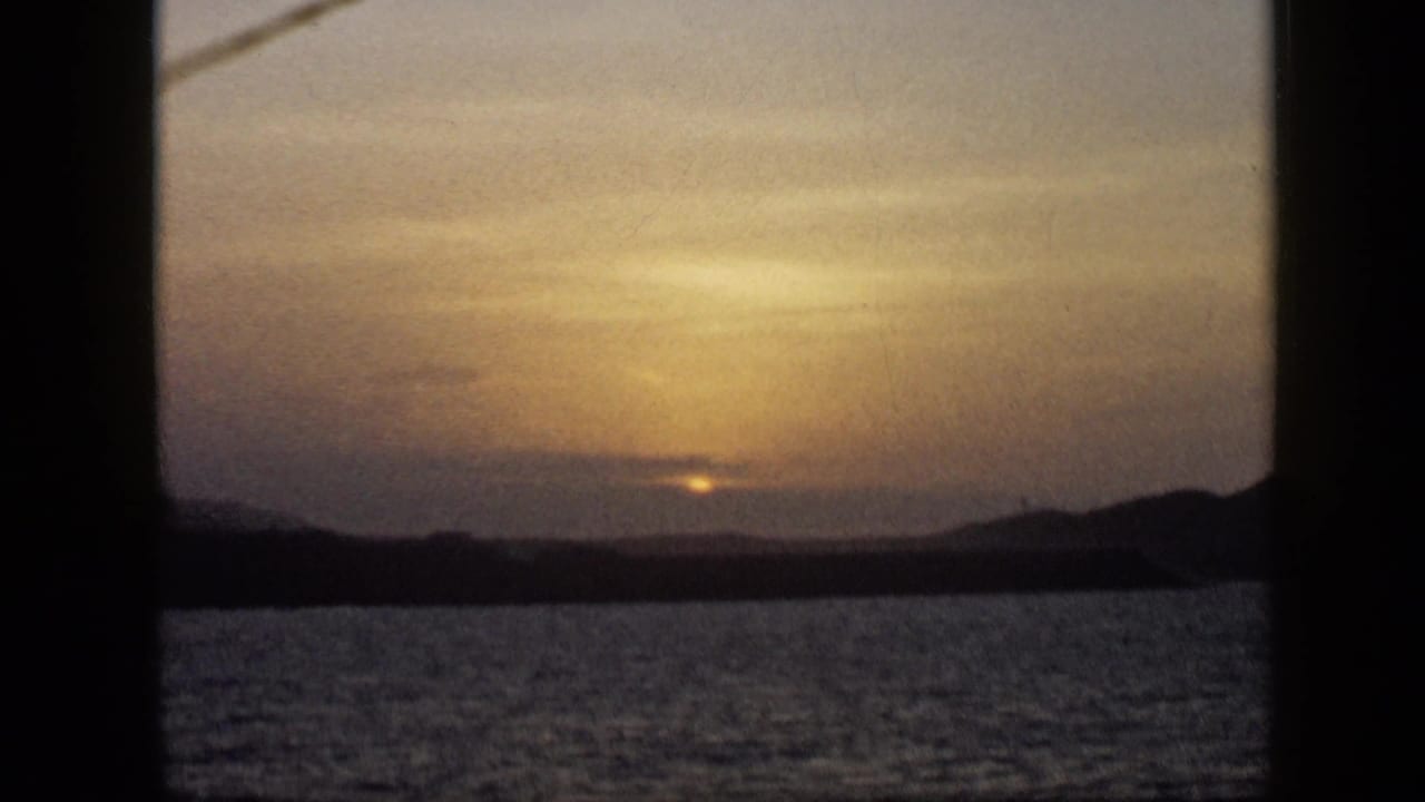 1959 Venezuela 8mm sunset scene showing a dim sun, calm sea, silhouetted hills, and dark framing bars around the view.