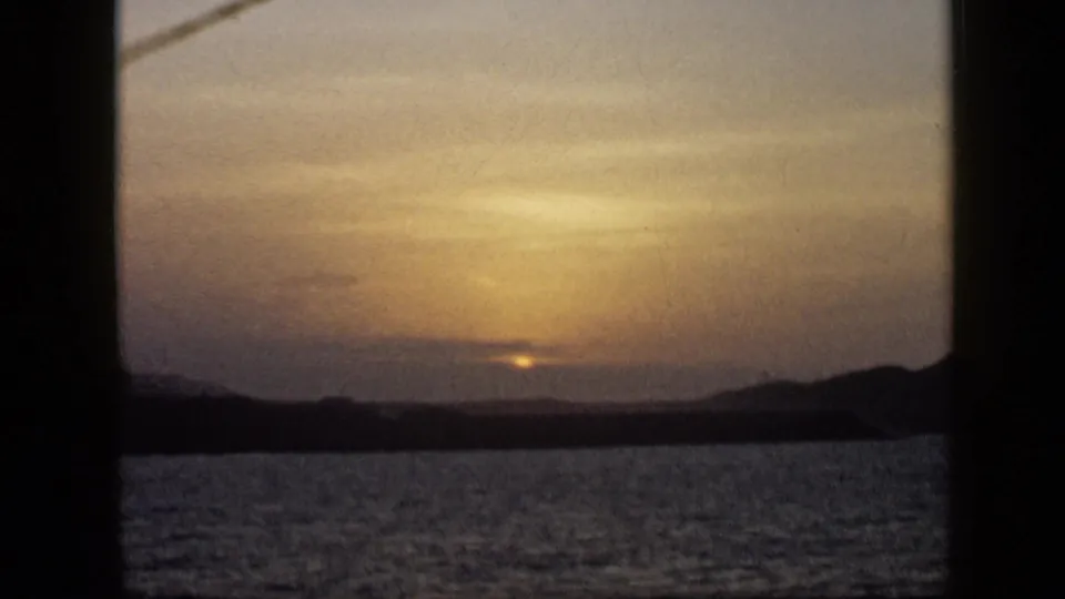 1959 Venezuela 8mm sunset scene showing a dim sun, calm sea, silhouetted hills, and dark framing bars around the view.