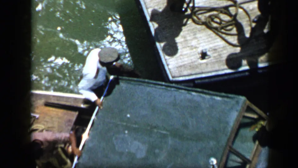 Man in a white shirt and hat boards or works beside a dock in Venezuela, pulling a large green crate near coiled rope.
