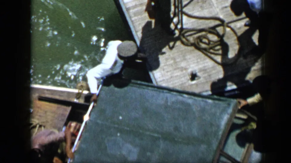 1959 Venezuela home-movie frame showing men on a wooden dock handling rope near a large metal crate beside green water.