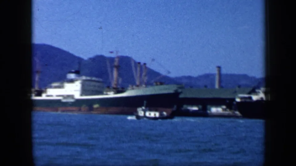 1959 Venezuela 8mm frame showing a docked cargo vessel, a small boat, harbor water, and a mountainous shoreline.