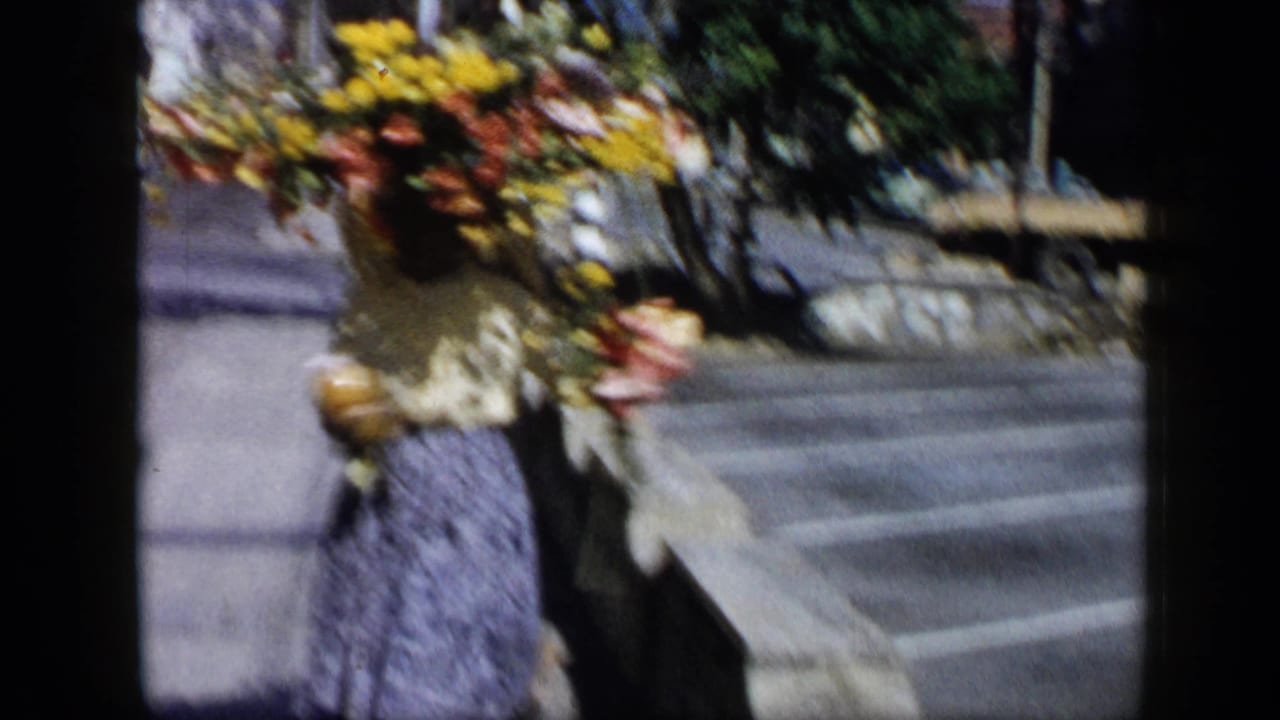 Woman walking on a sidewalk while carrying a large floral arrangement on her head.