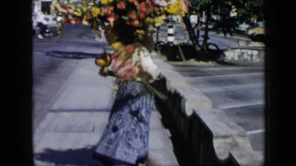 1959 Venezuela home-movie frame of a woman carrying flowers on her head near a low wall, sidewalk, street, trees, and parked cars.