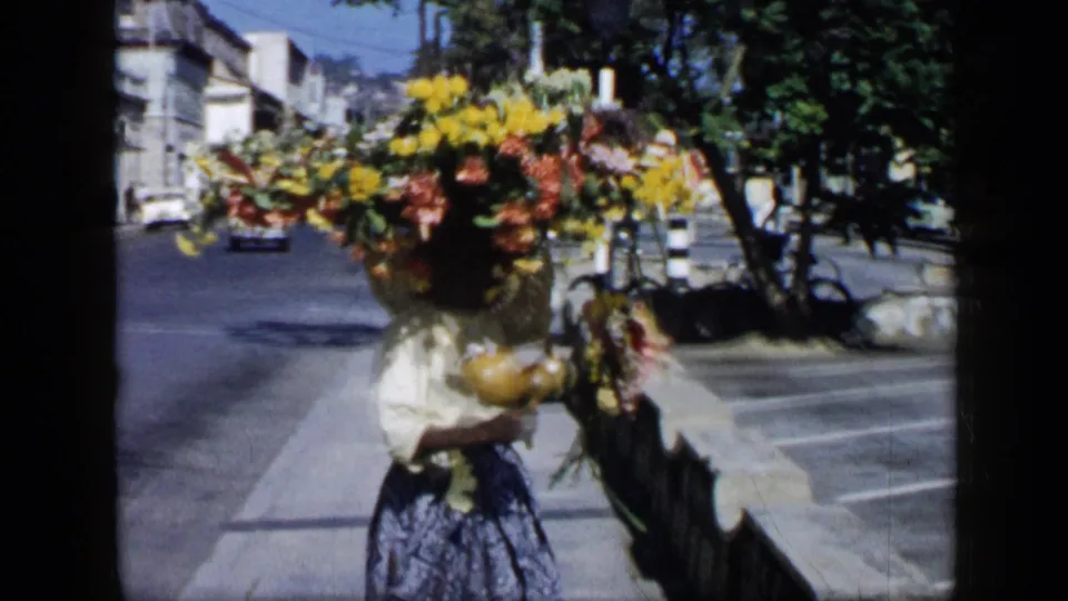 1959 Venezuela street scene showing a woman with a large flower arrangement on her head, walking near a curb, cars, trees, and buildings.