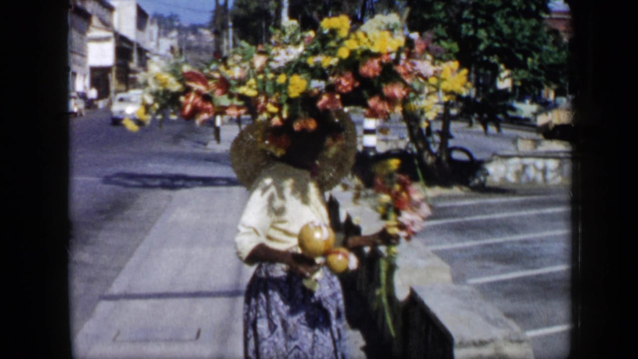 In Venezuela in the supplied 1959 context, a woman in a light blouse and patterned skirt carries a large floral arrangement on her head and holds maracas beside a city street.