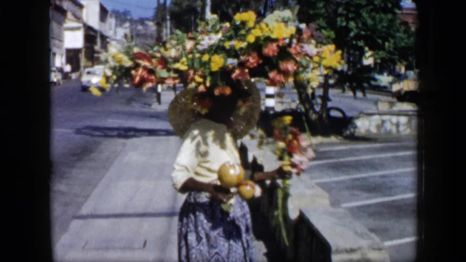 In Venezuela in the supplied 1959 context, a woman in a light blouse and patterned skirt carries a large floral arrangement on her head and holds maracas beside a city street.