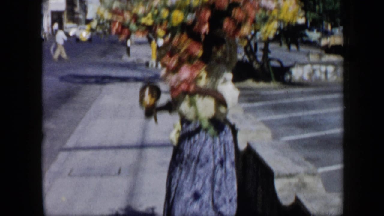 Woman under a large floral arrangement moves along the sidewalk with maracas in hand.