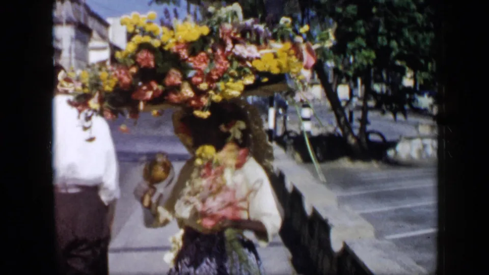 Near the end of the clip, the woman carrying flowers and maracas passes close to the camera on a bright Venezuelan city sidewalk, with another adult partly visible beside her.