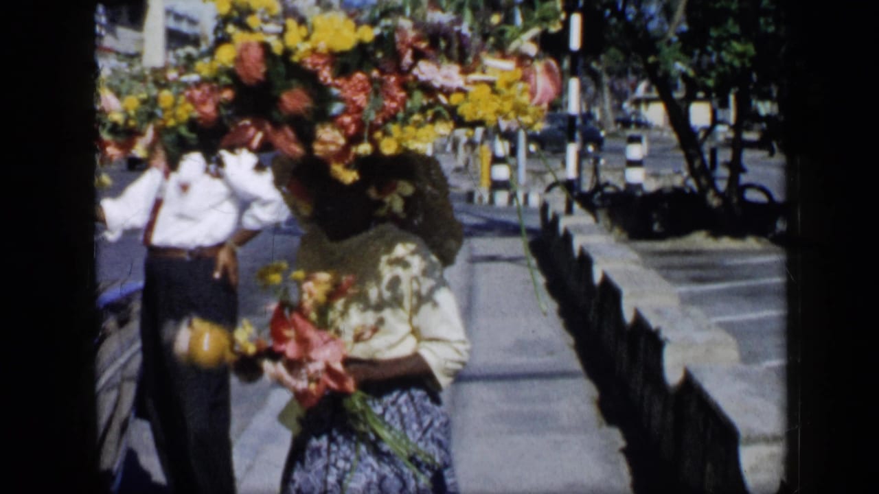 In 1959 Venezuela, a woman in a patterned skirt and flower crown carries flowers along a paved street beside a man holding yellow fruit.