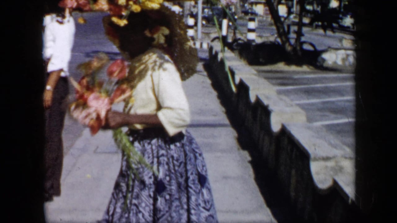 In 1959 Venezuela, a woman with flowers and a floral headpiece moves along a bright street with trees and low stone borders nearby.