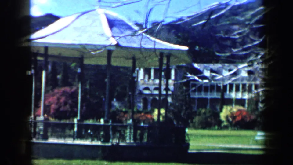 A 1959 Venezuela park scene frames a gazebo, clipped lawn, flowering shrubs, trees, distant mountains, and bright blue sky.