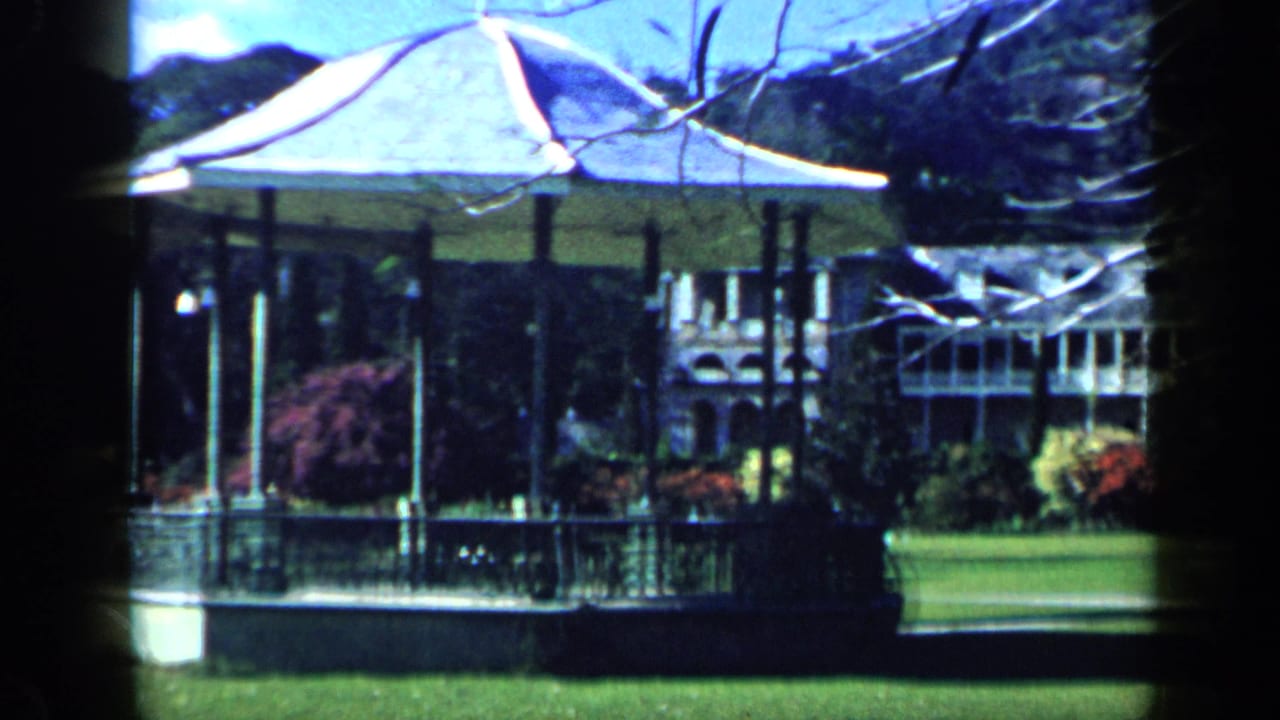 1959 Venezuela home-movie frame showing a white gazebo, lawn, trees, and a large estate building beyond.