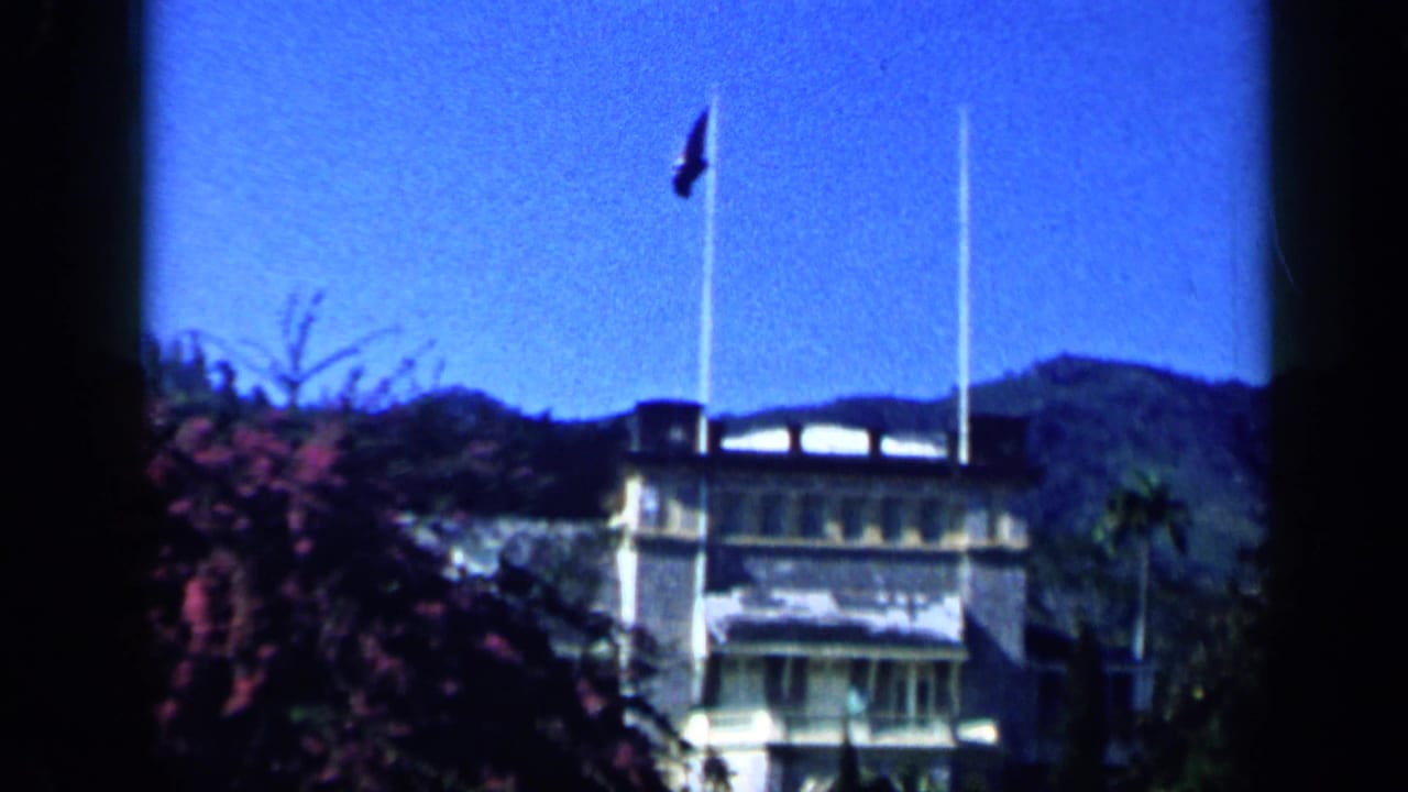 1959 Venezuela home-movie frame showing flagpoles, a low building, flowering shrubs, and distant hills.