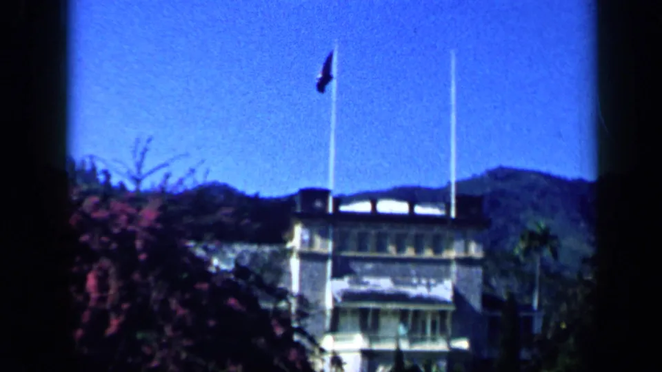 1959 Venezuela home-movie frame showing flagpoles, a low building, flowering shrubs, and distant hills.