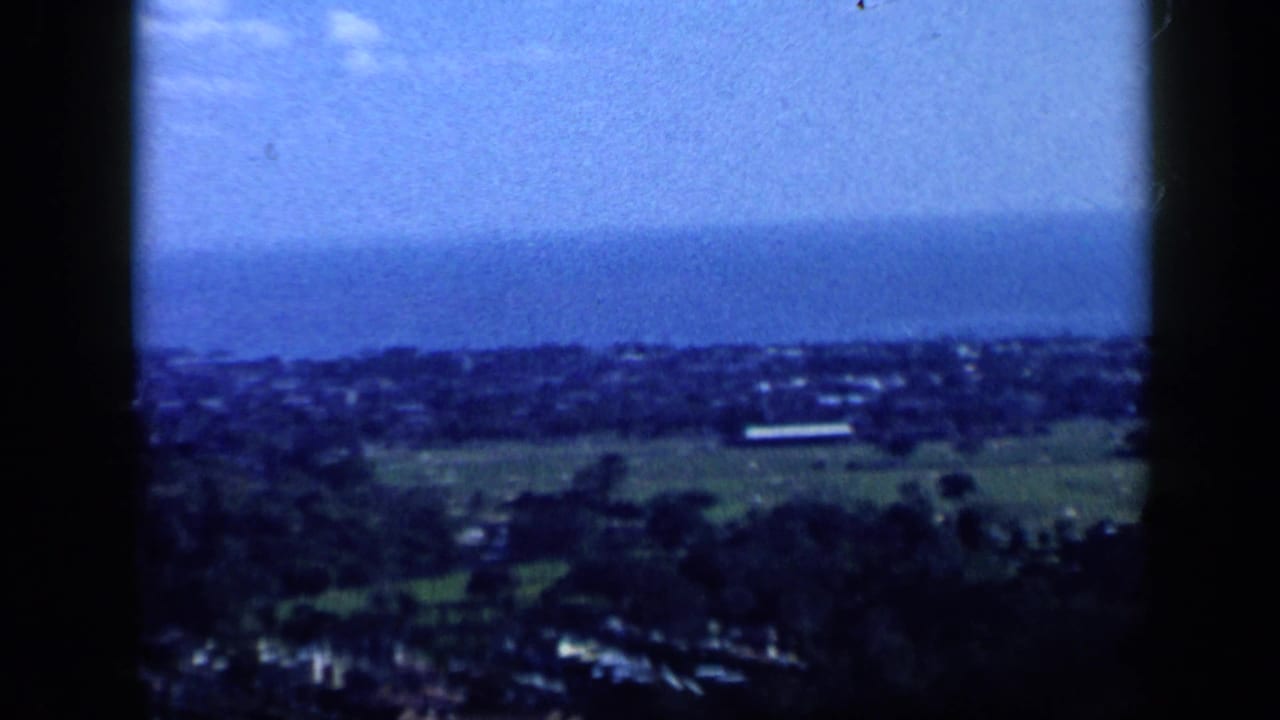 1959 Venezuela home-movie frame showing a broad hilltop view over coastal buildings, water, and distant landforms.
