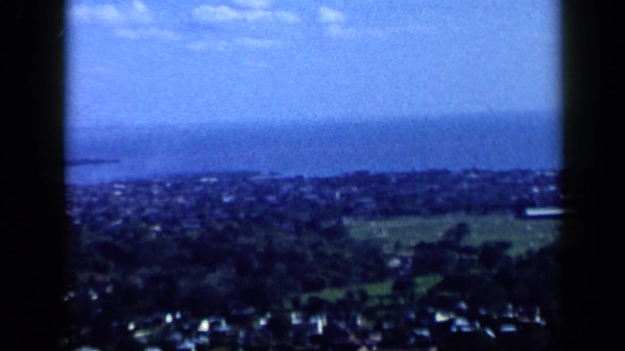 Soft aerial view of a coastal town with houses, hills, sea, and hazy sky.