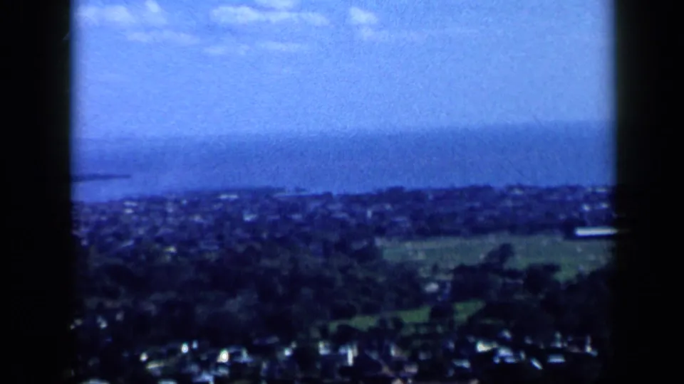 1959 Venezuela aerial home-movie frame showing a coastal town below green hills and a distant blue shoreline.