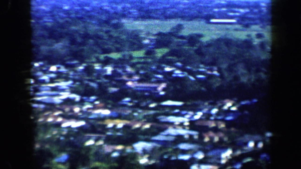 1959 Venezuela home-movie frame showing a dense neighborhood, green fields, hills, and a distant shoreline.