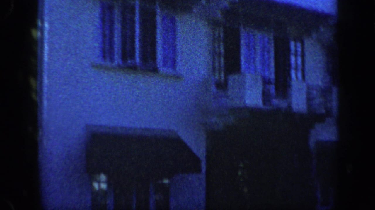 Blue-tinted 1959 Venezuela 8mm frame showing a dim residential stucco facade with windows, awnings, and soft focus.