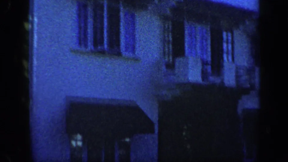 Blue-tinted 1959 Venezuela 8mm frame showing a dim residential stucco facade with windows, awnings, and soft focus.