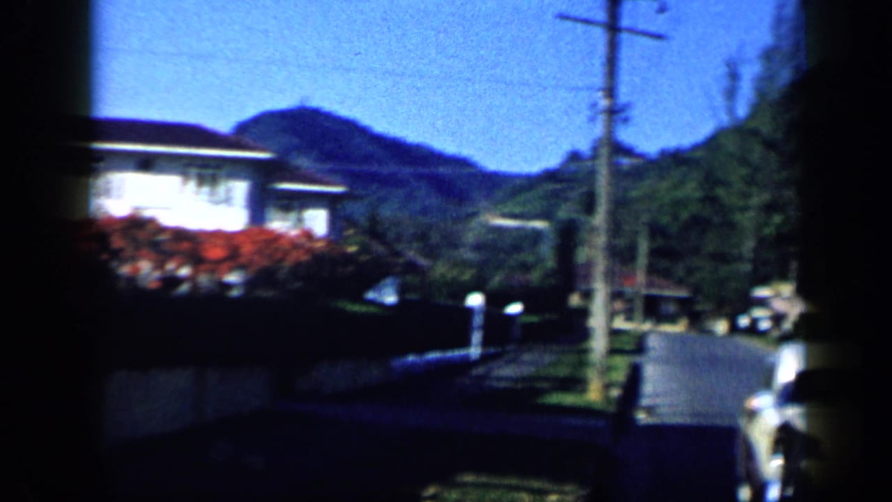 1959 Venezuela 8mm frame looking down a quiet residential street with a telephone pole, houses, gardens, hills, and parked cars.
