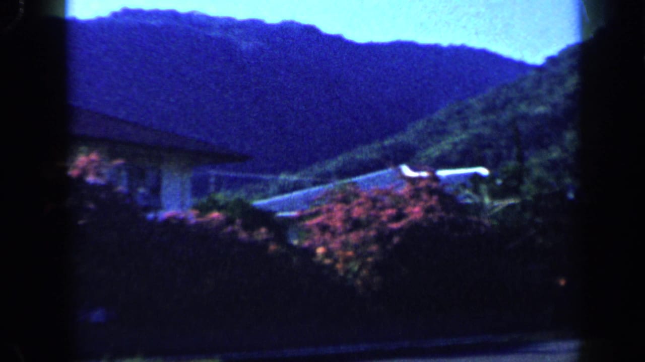 1959 Venezuela 8mm frame revealing a hillside residential view with greenery, a house roofline, and saturated blue mountain shadows.