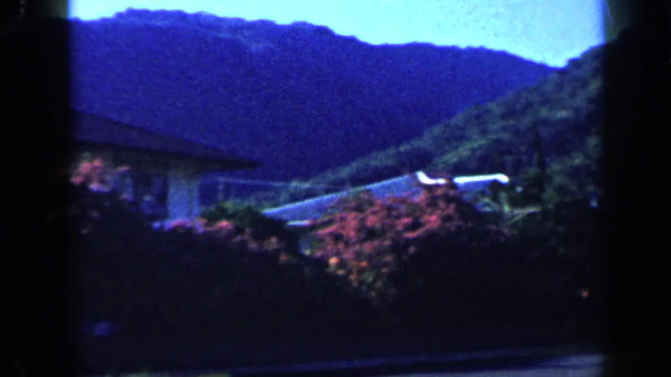 1959 Venezuela 8mm frame revealing a hillside residential view with greenery, a house roofline, and saturated blue mountain shadows.