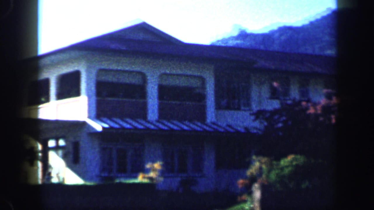 1959 Venezuela home-movie frame showing a two-story house, large windows, garden growth, and distant hillside through shaded foreground edges.