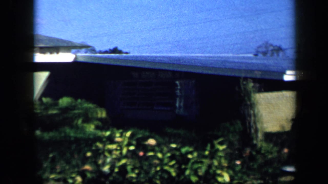 1959 Venezuela exterior frame showing a single-story house with a low roof, shuttered window, garden plants, and bright blue sky.