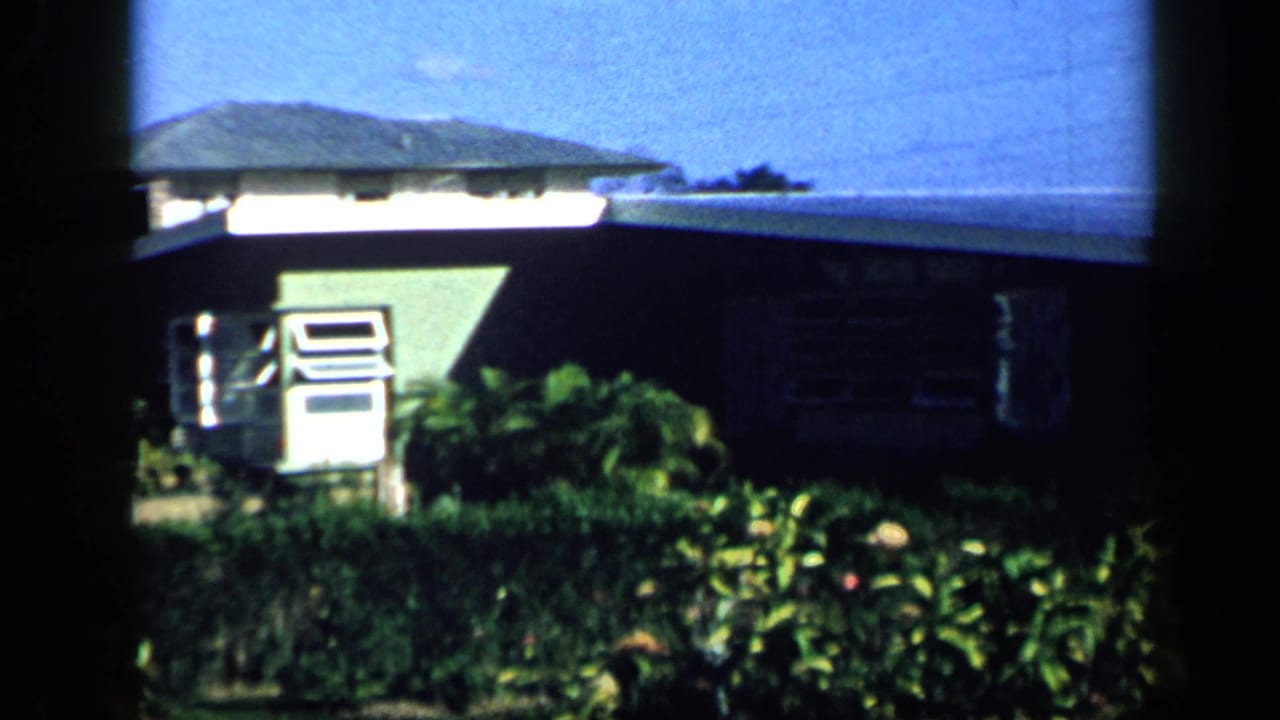 1959 Venezuela 8mm frame showing a green residential exterior with a low roofline, louvered window, garden plants, and blue sky.