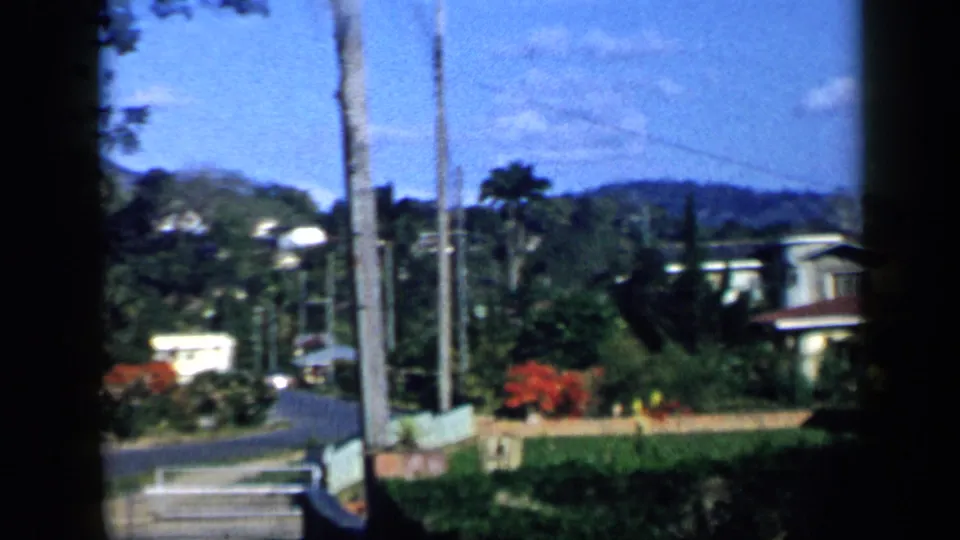 1959 Venezuela 8mm frame showing a residential street area with palm trunks, garden walls, houses, utility lines, and distant hills.