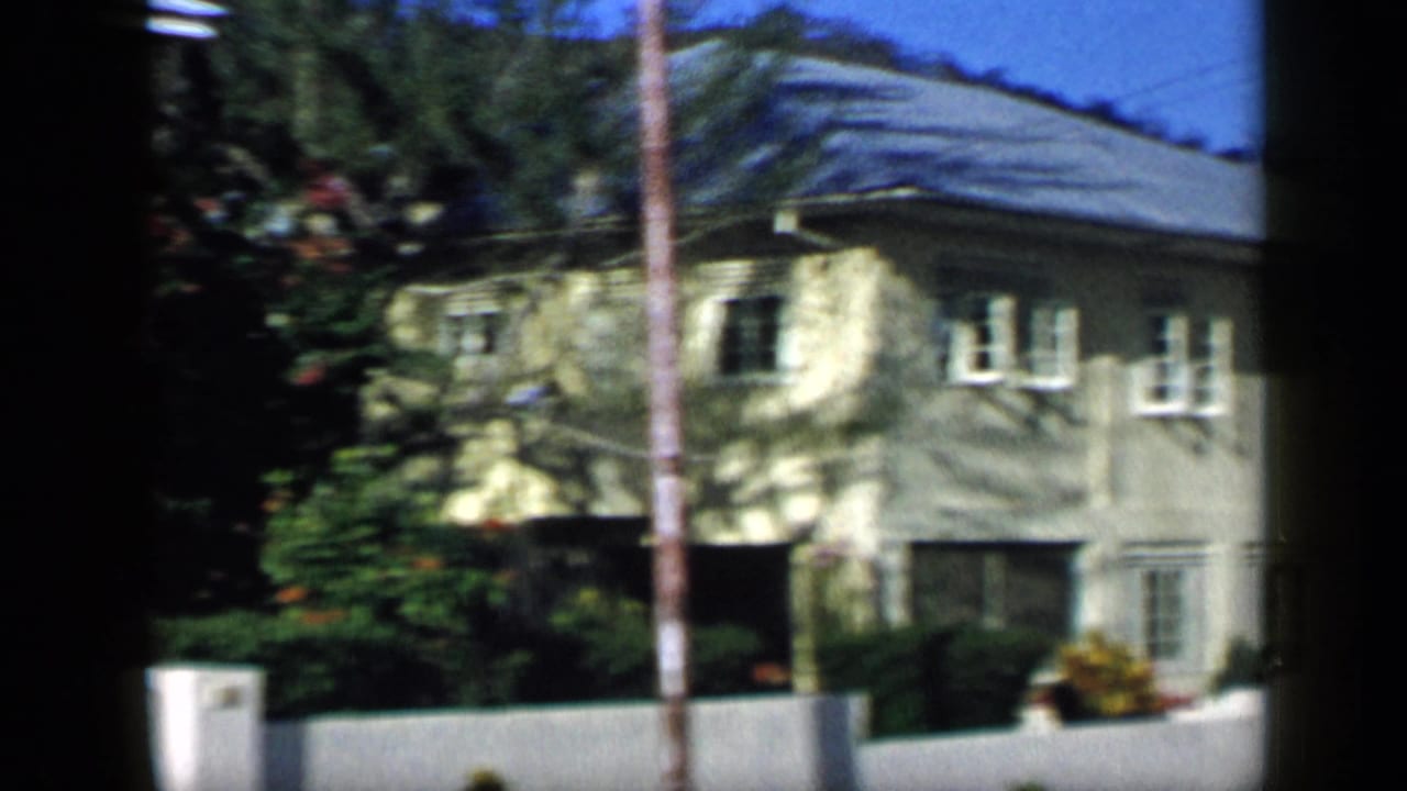 1959 Venezuela 8mm frame showing a two-story residential building with white shutters, a pale facade, palm shadow, garden plants, and a low wall.
