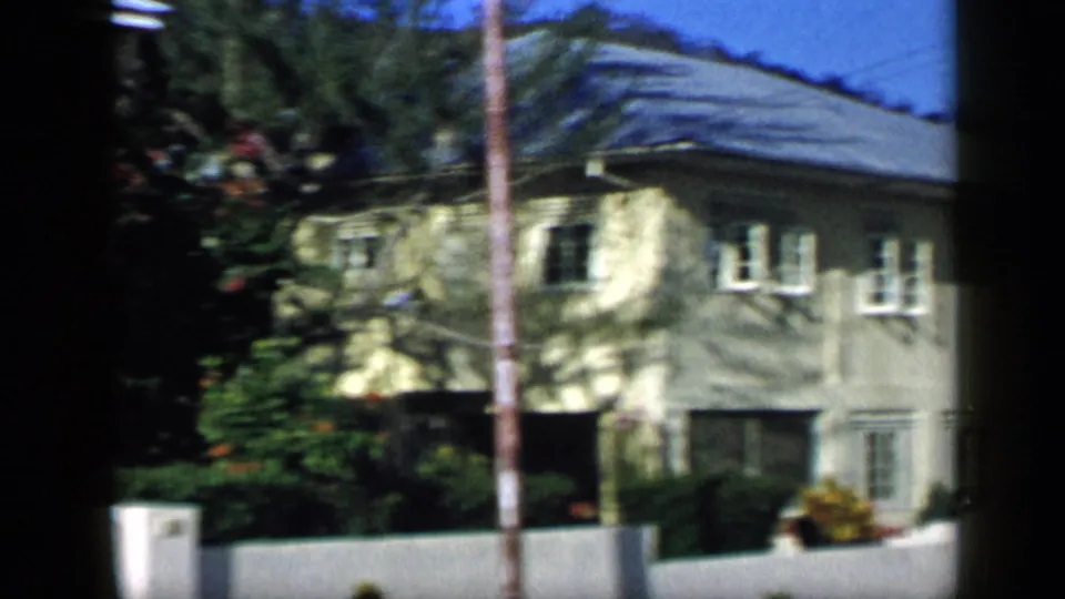 1959 Venezuela 8mm frame showing a two-story residential building with white shutters, a pale facade, palm shadow, garden plants, and a low wall.