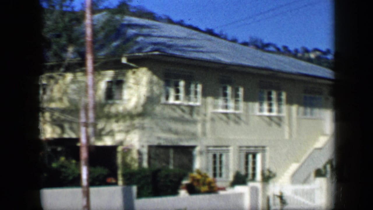 1959 Venezuela 8mm frame showing a light-colored house with white-trimmed windows, a fence, palms, and green hillside beyond.
