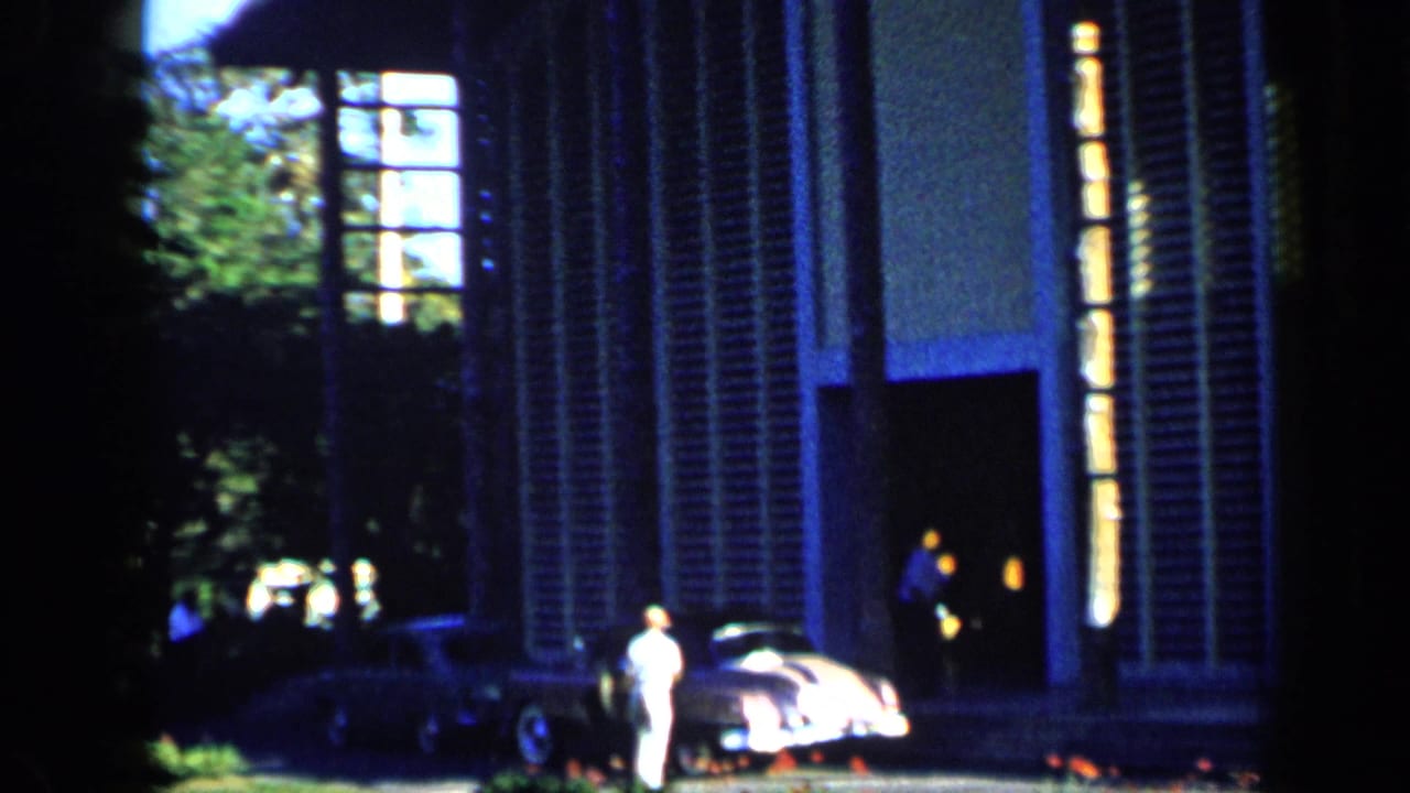 1959 Venezuela frame showing a shuttered house facade, a parked rounded car, and people near the entrance in deep shade.