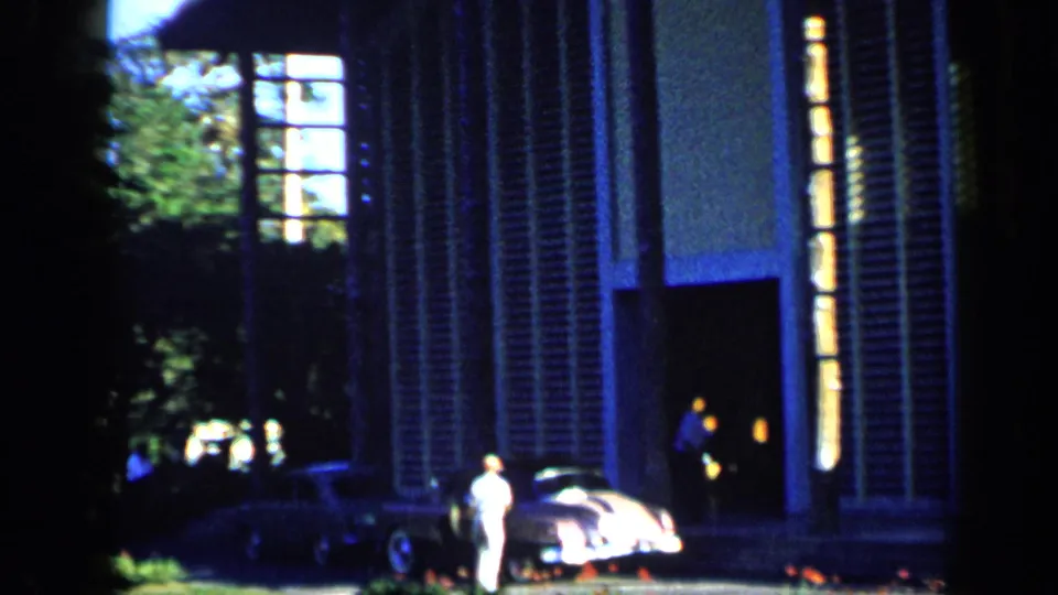 1959 Venezuela frame showing a shuttered house facade, a parked rounded car, and people near the entrance in deep shade.