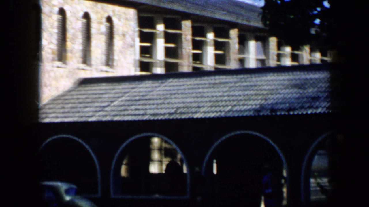 1959 Venezuela exterior frame with arched walkways, roof tiles, upper windows, and a partly obscured vehicle near the shaded arcade.