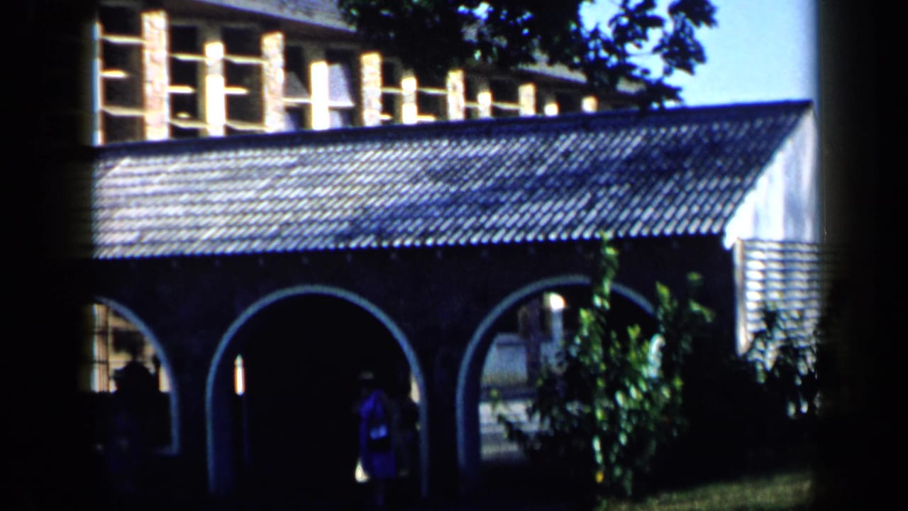 1959 Venezuela frame showing the building's arcade, tile roof, greenery, lawn, and distant figures moving in shadow.