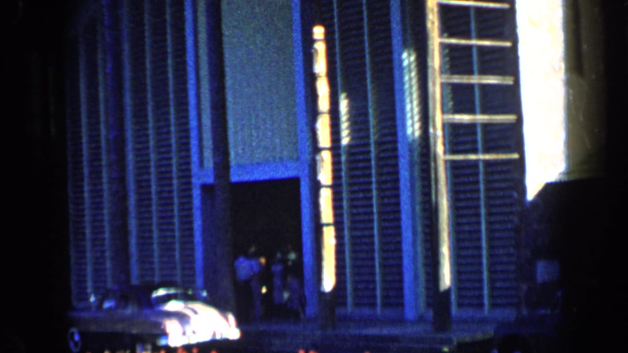 1959 Venezuela 8mm frame showing a modern facade with vertical blue-tinted panels, glowing lights, a car, and distant people.