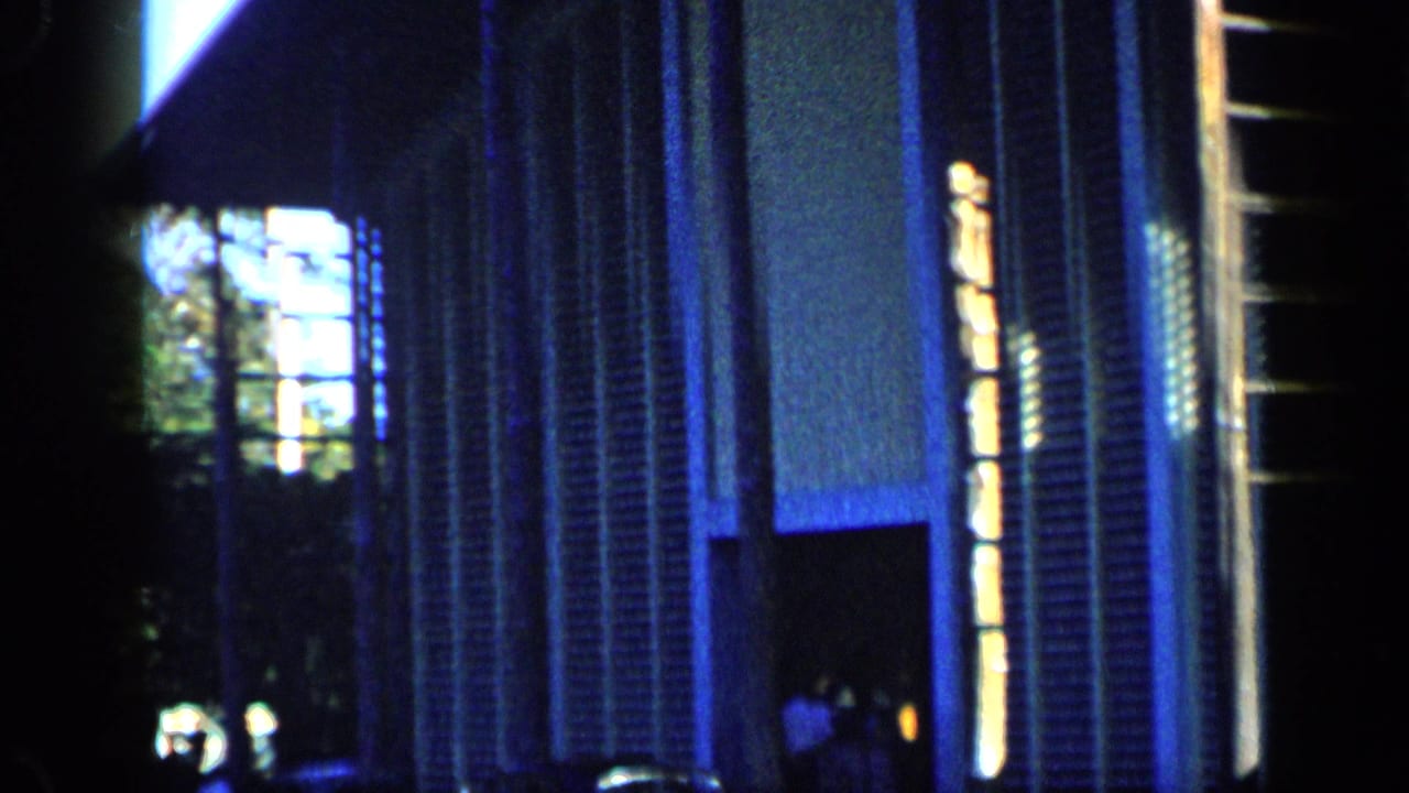 1959 Venezuela frame with a dark, blue-tinted modern building exterior, bright vertical lighting, and small people near the doorway.