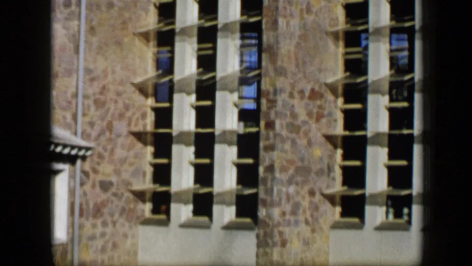 1959 Venezuela 8mm frame showing a stone facade with white vertical window frames and shadowed multi-paned openings.