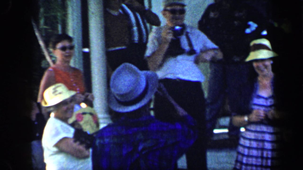 1959 Venezuela home-movie frame showing people in hats and summer clothing gathered outside near a shaded structure.