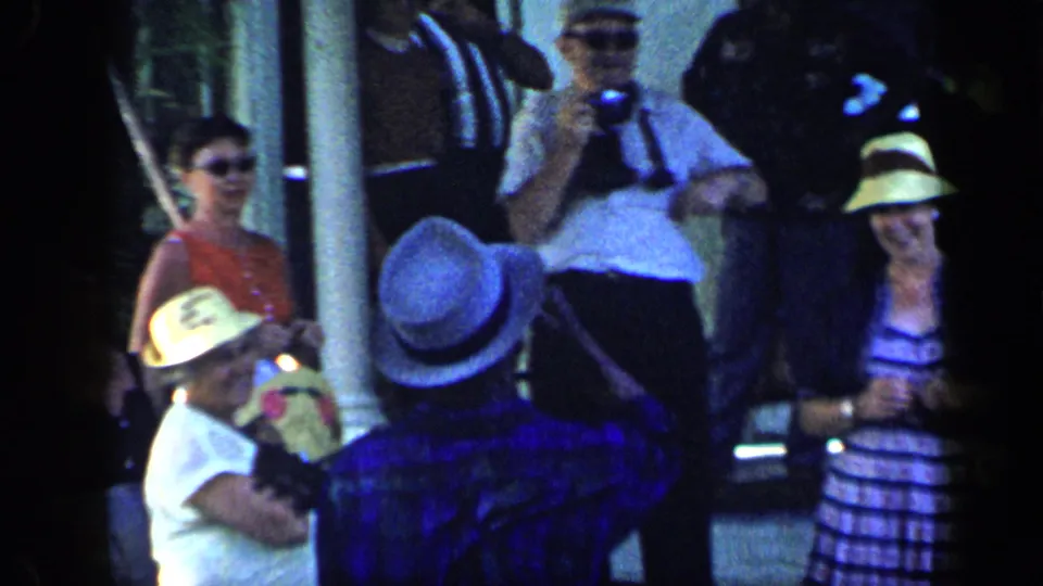 1959 Venezuela home-movie frame showing people in hats and summer clothing gathered outside near a shaded structure.