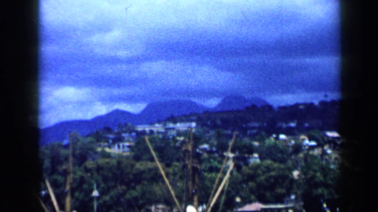 Mountainous coastal city with harbor masts and cranes under a cloudy sky.