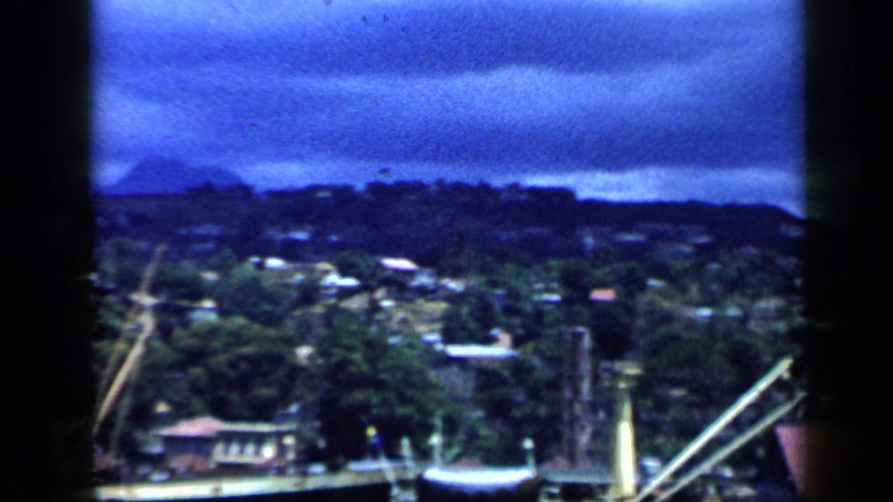 1959 Venezuela 8mm view of a mountainous urban waterfront with cranes, clustered buildings, and heavy blue cloud cover.